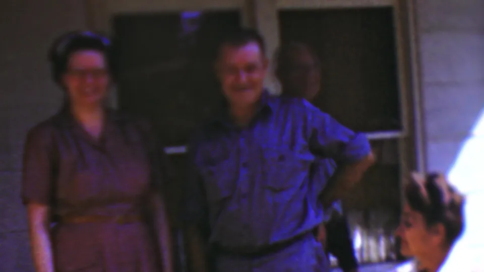 1954 Wisconsin home-movie frame of a woman in a brown dress and a man in a blue shirt smiling on a porch, with another adult standing in shadow and a seated woman partly visible.