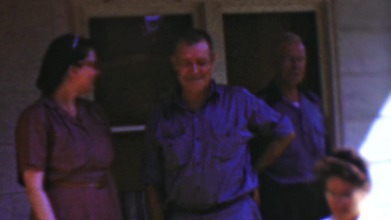 In this 1954 Wisconsin home-movie frame, adults stand on or near a porch as a woman in a purple dress faces a smiling man in a blue shirt, with another man partly visible in shadow.