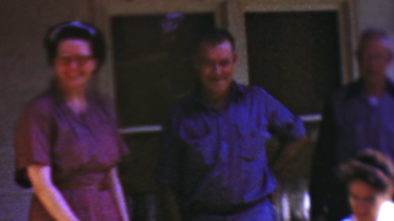 A woman in purple and a smiling man in blue stand in front of screened doors while another adult is partly visible.