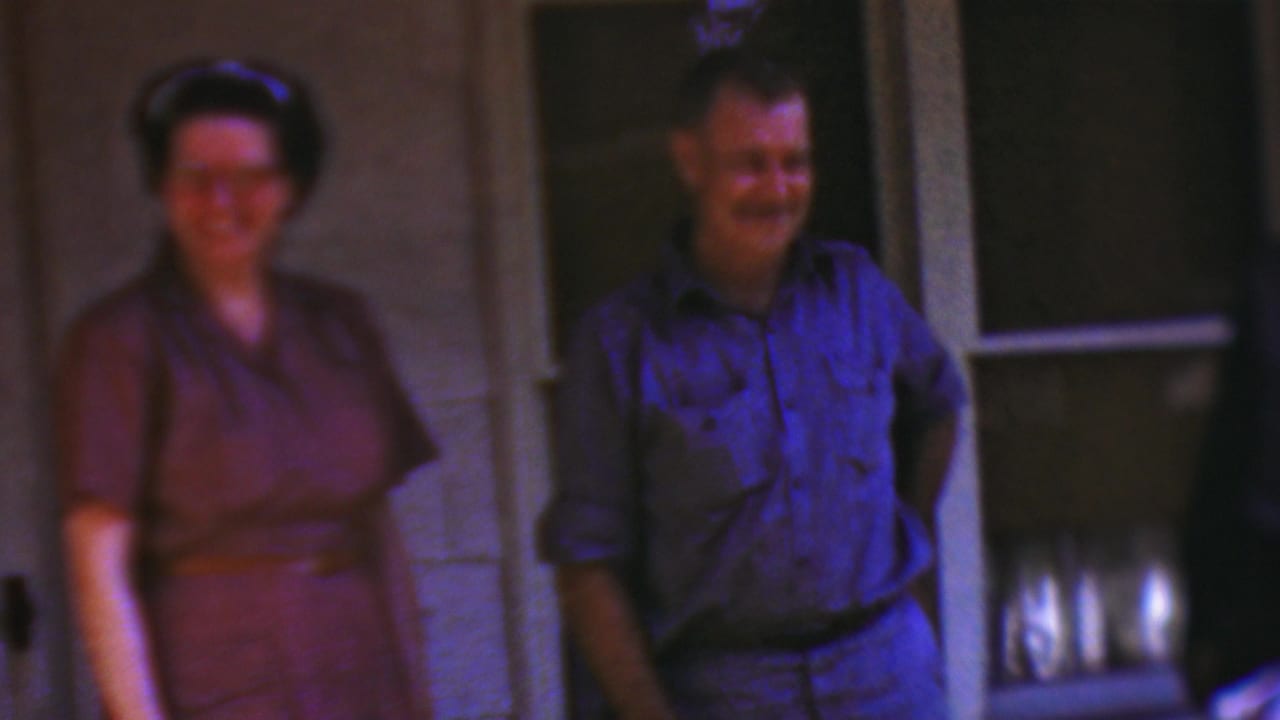 In the 1954 Wisconsin porch scene, a woman in a purple dress stands beside a smiling man in a blue shirt, with the house wall and dark screened doorway behind them.