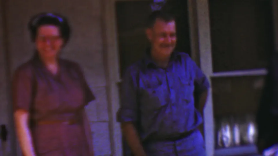 In the 1954 Wisconsin porch scene, a woman in a purple dress stands beside a smiling man in a blue shirt, with the house wall and dark screened doorway behind them.