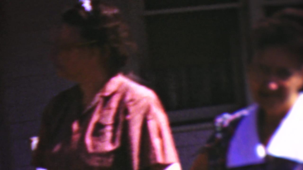 In metadata-supported 1954 Wisconsin home-movie footage, two adults stand close to a building facade, with one person in a pink collared shirt and glasses partly turned toward the camera.