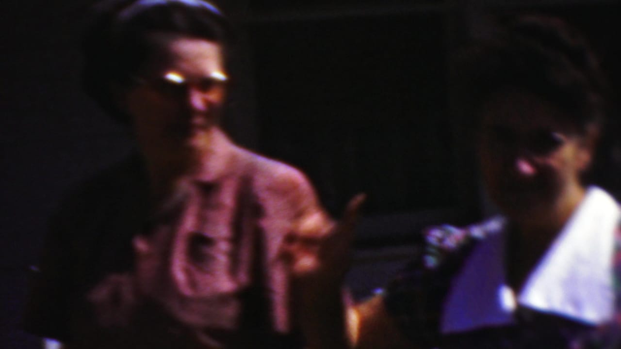 Two women in mid-century clothing smile toward the camera in the 1954 Wisconsin home-movie scene, with a man in darker clothing standing between them in partial shadow.