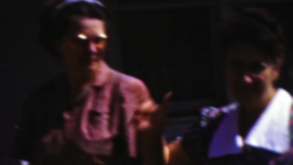Two women in mid-century clothing smile toward the camera in the 1954 Wisconsin home-movie scene, with a man in darker clothing standing between them in partial shadow.