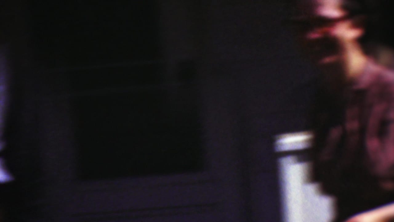 A handheld 8mm frame shows a blurred adult in a dark shirt near a building exterior, with another person only partly visible along the left edge.