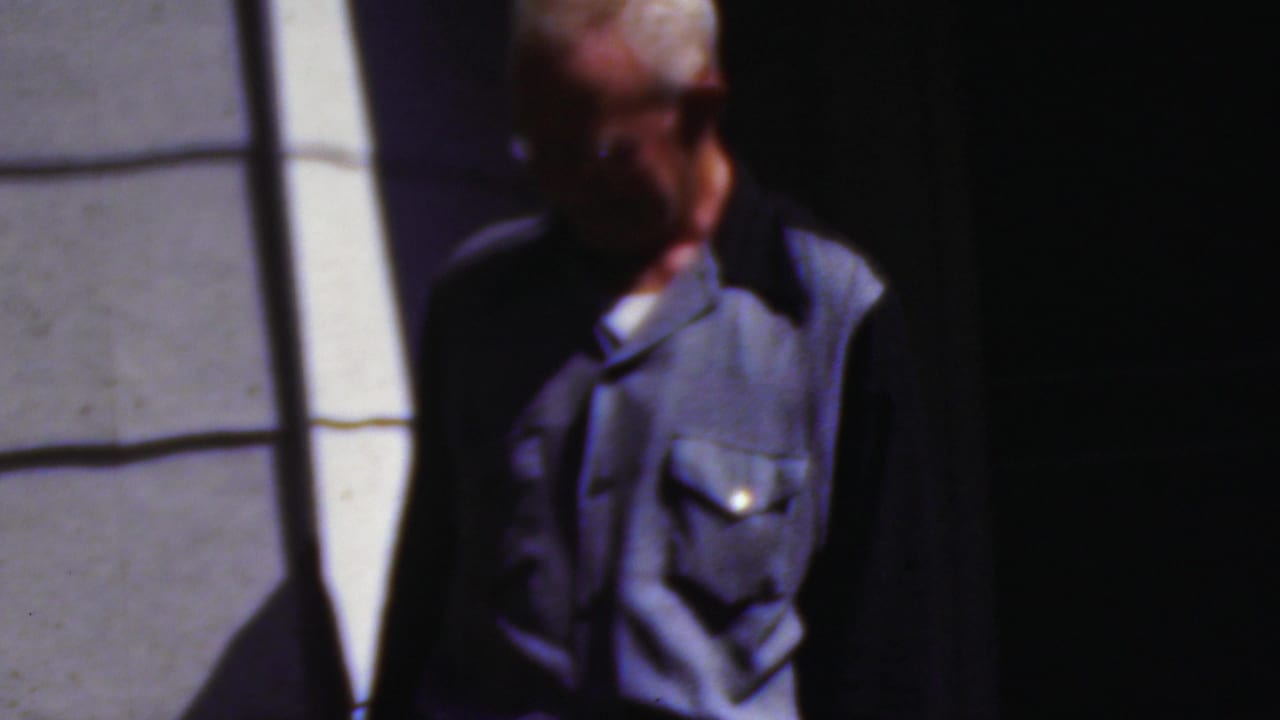 An older adult wearing a blue shirt or vest stands beside a dark doorway or entrance in the metadata-supported 1954 Wisconsin home-movie footage.