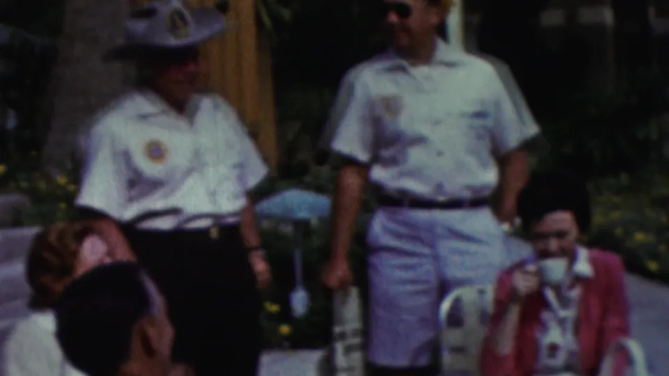 California 1961 home-movie frame showing several men at an outdoor gathering, with two standing in light shirts with visible patches and seated attendees in the foreground.