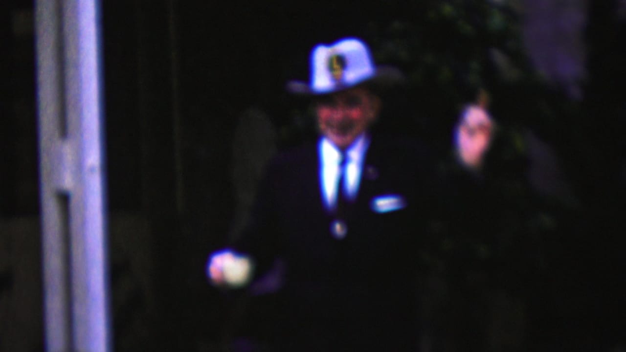 Man in a white cowboy-style hat and dark suit walks outdoors while holding a white cup and raising one hand.