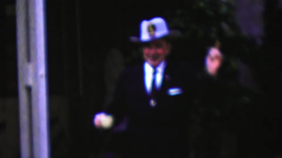 California 1961 frame of a smiling man in a white cowboy-style hat and dark suit walking through a dim outdoor setting with a white cup in one hand.