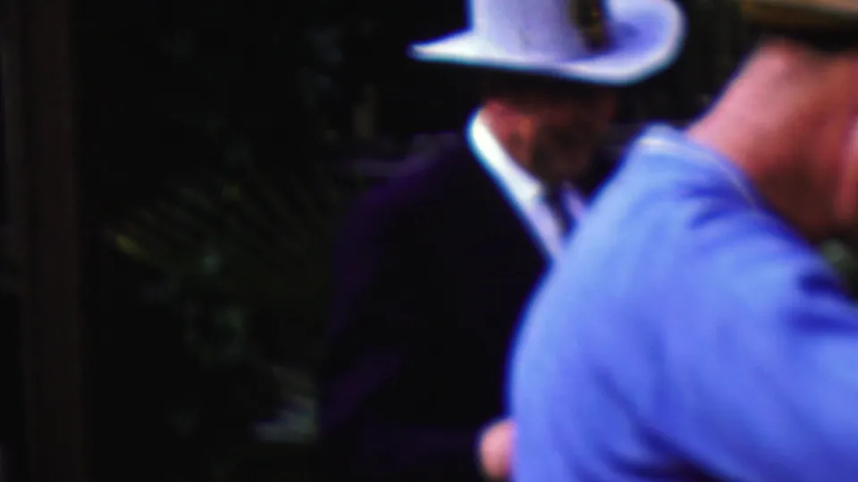 California 1961 frame showing the white-hatted man in a dark suit moving past the camera area, with another attendee in a light blue shirt partly visible in the foreground.