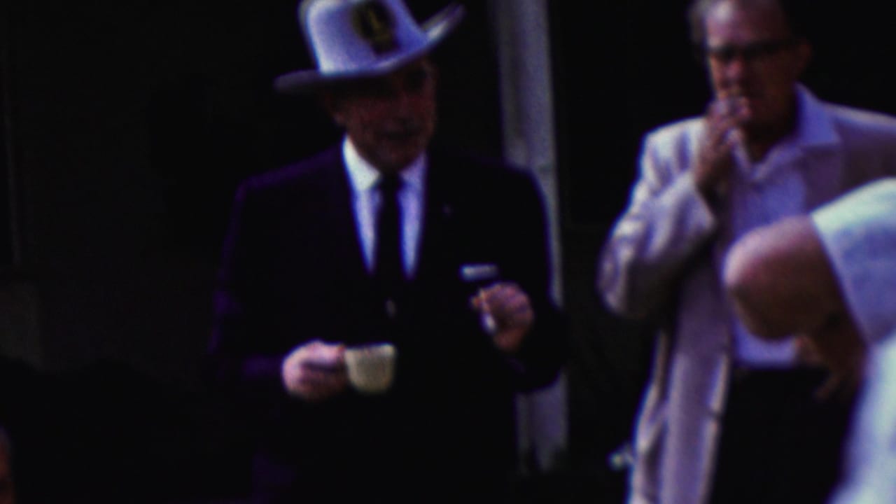 In this 1961 California home-movie frame, a man in a dark suit and white cowboy hat holds a small cup as men in light clothing stand nearby in a shaded outdoor setting.