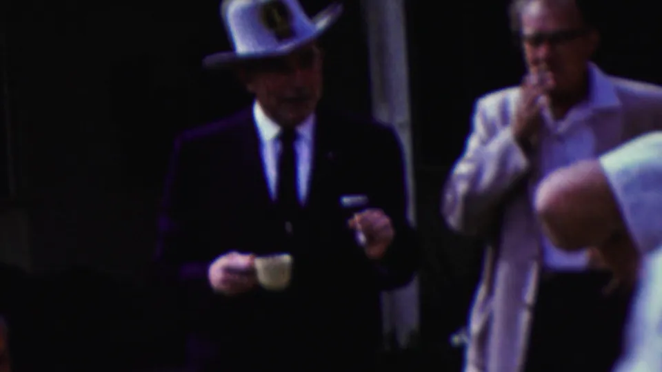 In this 1961 California home-movie frame, a man in a dark suit and white cowboy hat holds a small cup as men in light clothing stand nearby in a shaded outdoor setting.