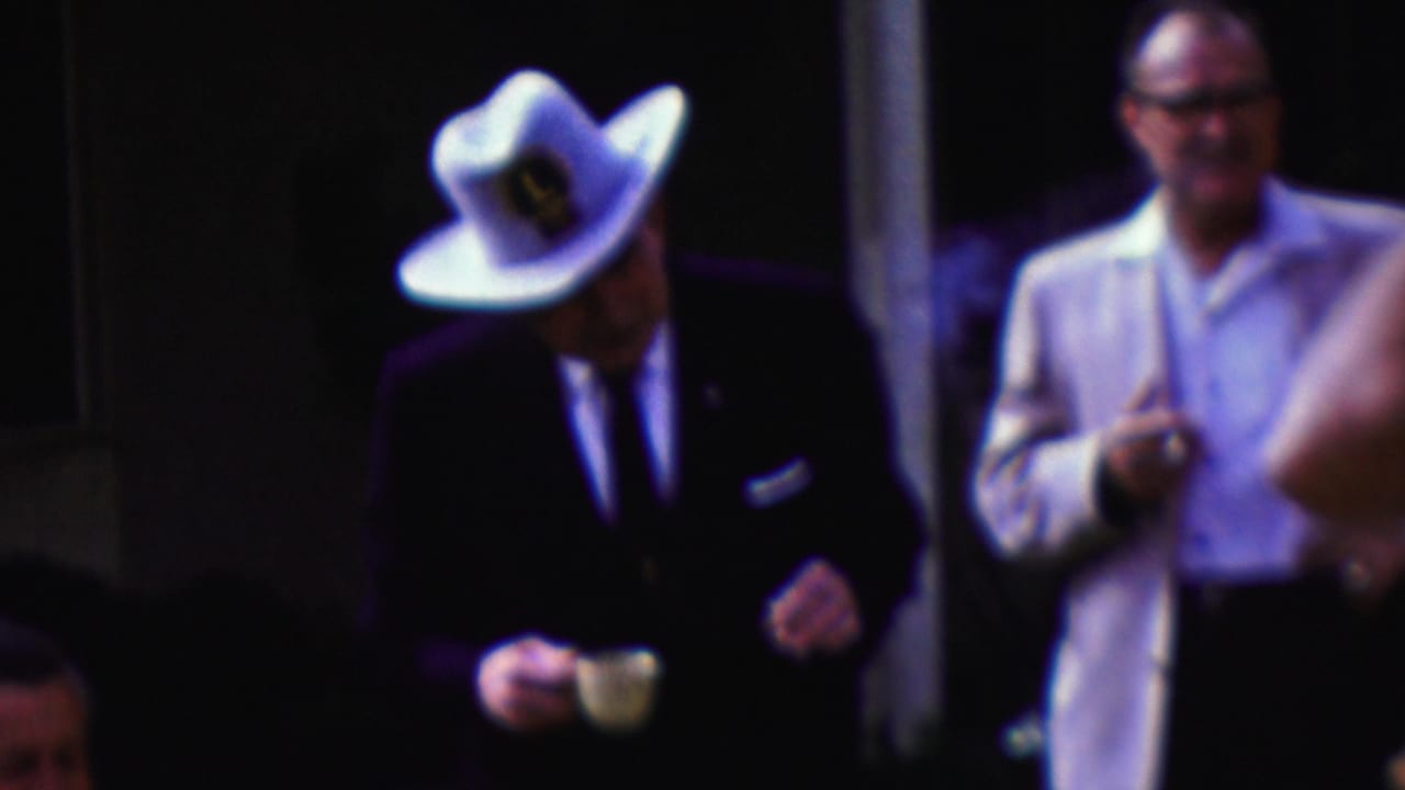 The frame shows the suited man in a white cowboy hat looking downward with a small cup in hand, with other adult men standing close by in the dim California gathering scene.