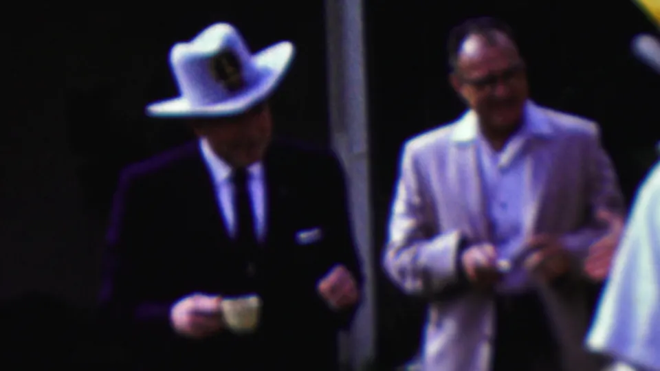 A man in a dark suit and white cowboy hat stands with a small cup while a man in a light jacket and glasses smiles nearby, suggesting informal conversation within the 1961 California gathering.