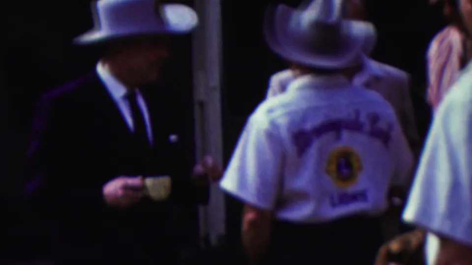 1961 California home-movie frame showing adults at a crowded outdoor Lions Club gathering, with a suited man holding a cup and a club member in a white cowboy hat seen from behind.