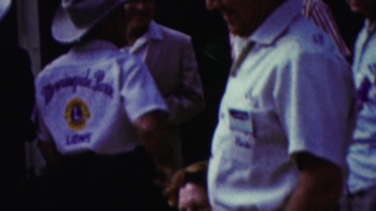 Several adults stand close together, including men in white shirts and cowboy hats with Lions Club markings.