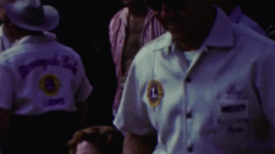 1961 California home-movie frame showing a close view of a man in a white Lions Club shirt, with another club member and adults in semi-formal clothing behind him.
