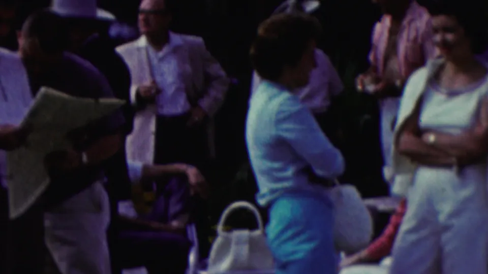 1961 California home-movie frame showing adults conversing at an outdoor event, with a man holding papers, a seated person partly visible, and a woman standing with folded arms.