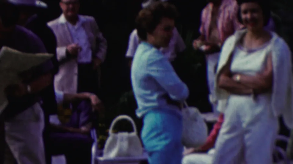 1961 California home-movie frame showing adults gathered outdoors, including a woman in a light blue sweater and turquoise pants near white chairs and handbags.