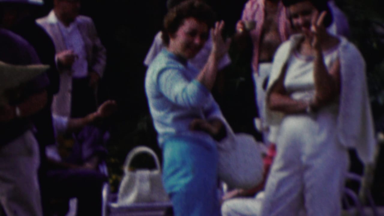 1961 California home-movie frame with a woman waving toward the camera during an outdoor gathering as other adults stand nearby in summer clothing.