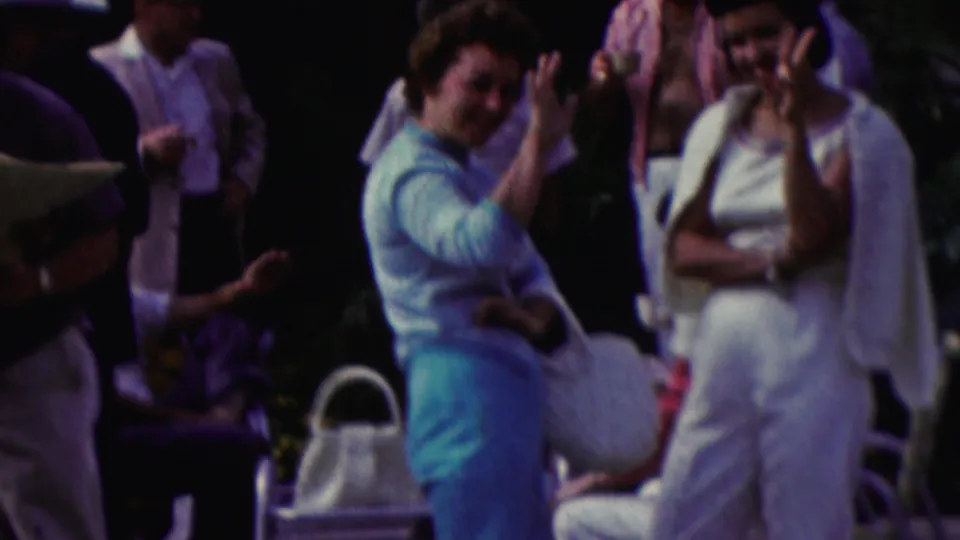 1961 California home-movie frame with a woman waving toward the camera during an outdoor gathering as other adults stand nearby in summer clothing.