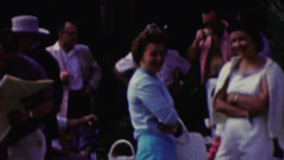 1961 California home-movie frame showing adults clustered near white chairs, including two women facing the camera and a partially visible capped figure at the edge.