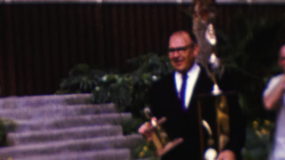 1961 California home-movie frame showing a man in a dark suit on stone steps holding a trophy-like object, with plants and other attendees nearby.
