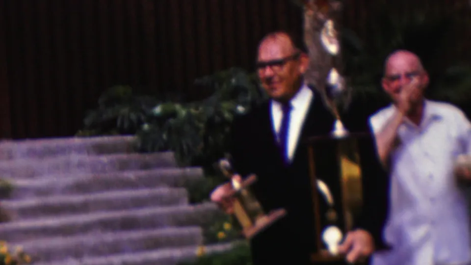 In this 1961 California home-movie frame, a suited man stands on landscaped stone steps holding a small trophy and a taller award while another man in a light shirt stands beside him.