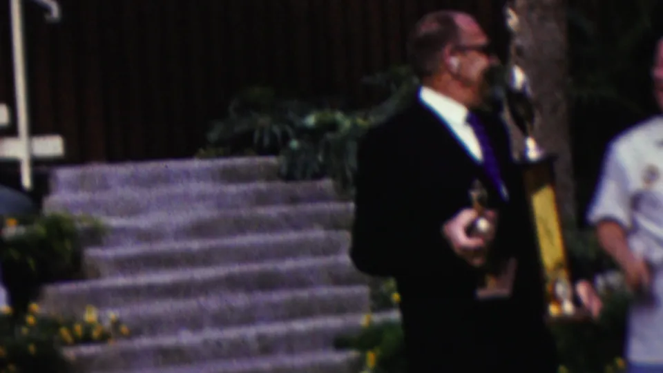 A suited man holding two awards turns in profile near stone steps, with landscaped plants and a partially visible adult in a light shirt behind him.