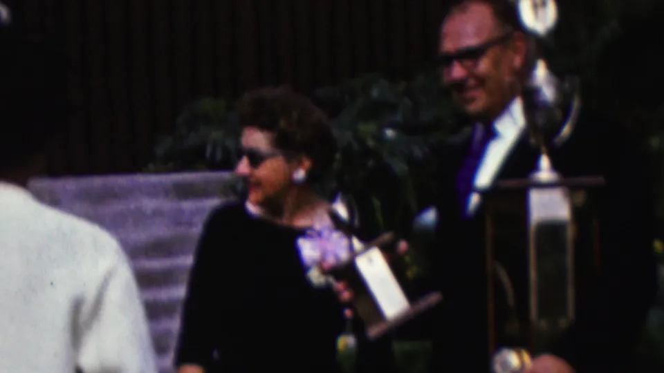 A woman in sunglasses and a dark outfit stands near a suited man holding a tall award, with stone steps and greenery behind them in the 1961 California home-movie scene.