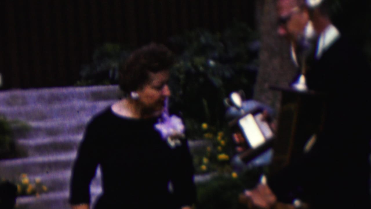 In California 1961 home-movie footage, a woman in a dark dress with a corsage stands beside stone steps while a suited man holds a large trophy nearby.