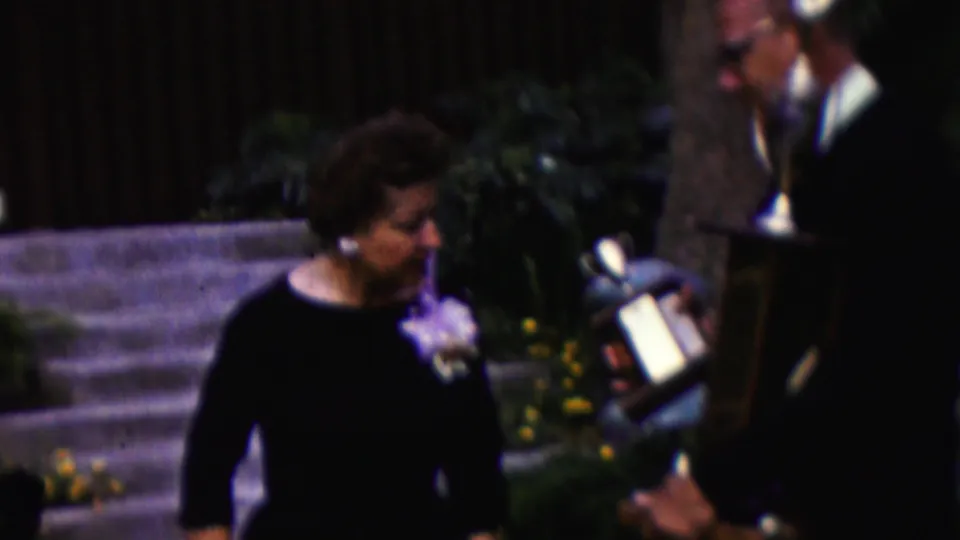 In California 1961 home-movie footage, a woman in a dark dress with a corsage stands beside stone steps while a suited man holds a large trophy nearby.