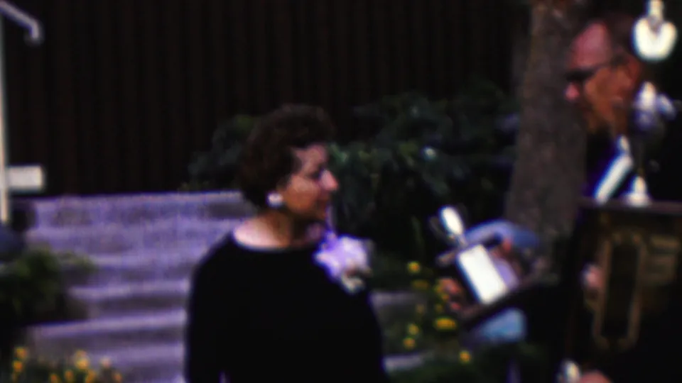 The woman turns toward the presented trophy during an outdoor California ceremony, with steps, shrubs, flowers, and a dark fence or wall behind her.