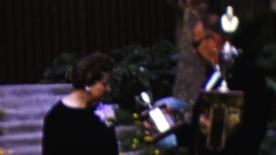 The woman smiles and leans slightly as the trophy is held close beside her, with the suited presenter, steps, planted flowers, and shrubs visible in the 1961 California scene.