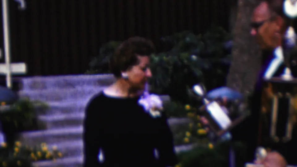 At the end of the clip, the woman stands beside the large trophy during the outdoor presentation, with the presenter partly visible and planted greenery behind them.