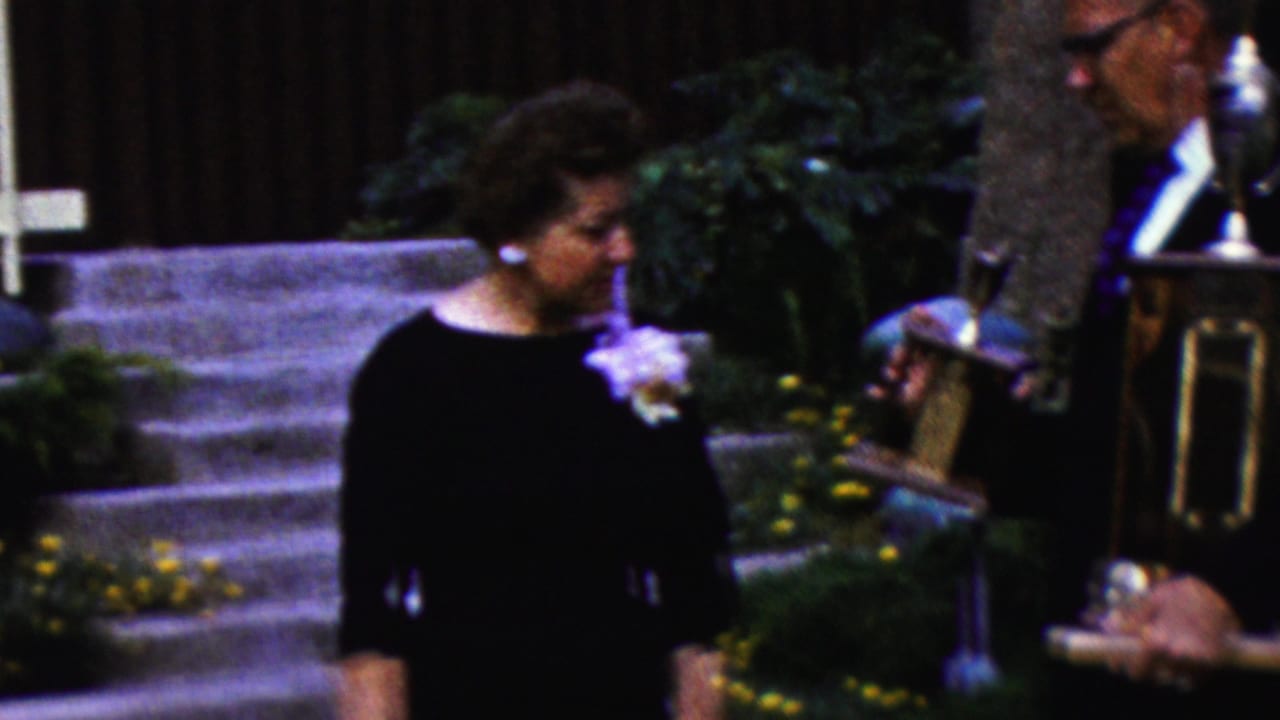 In 1961 California home-movie footage, an older woman wearing a dark dress and white corsage stands beside an older man holding a trophy near stone steps and garden plants.