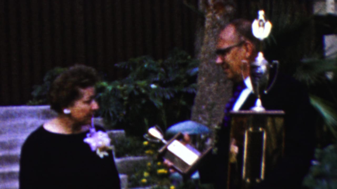 The older woman turns toward the older man as he presents or shows a smaller award object beside a larger trophy on a stand in the outdoor California ceremony scene.