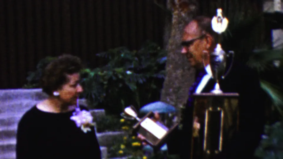 The older woman turns toward the older man as he presents or shows a smaller award object beside a larger trophy on a stand in the outdoor California ceremony scene.