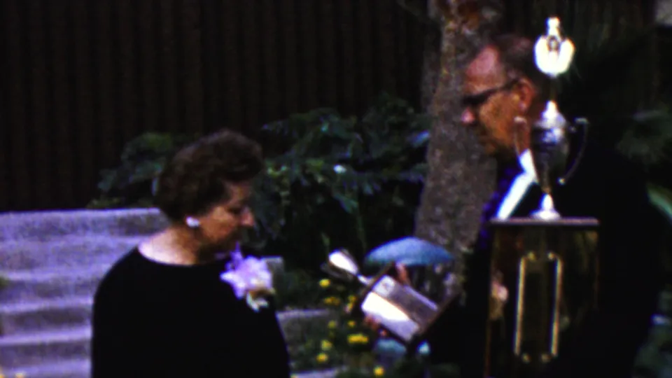 In the 1961 outdoor home-movie scene, the older woman looks toward a smaller trophy or plaque while the older man stands partly behind a large trophy on a stand.