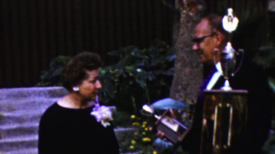 The final frame shows the older woman smiling toward the older man as he holds a small award object next to the prominent trophy stand in the outdoor 1961 California ceremony.