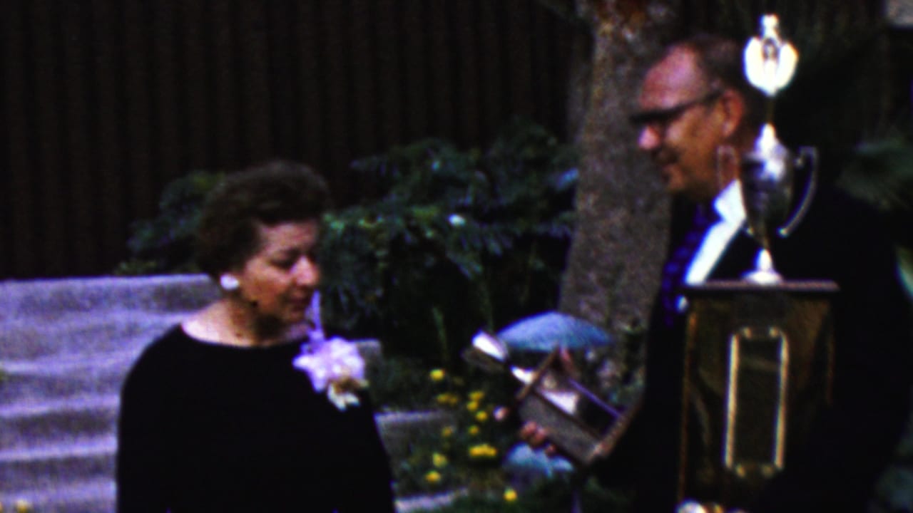 Woman in a black dress with a corsage stands beside a suited man holding a large trophy outdoors.