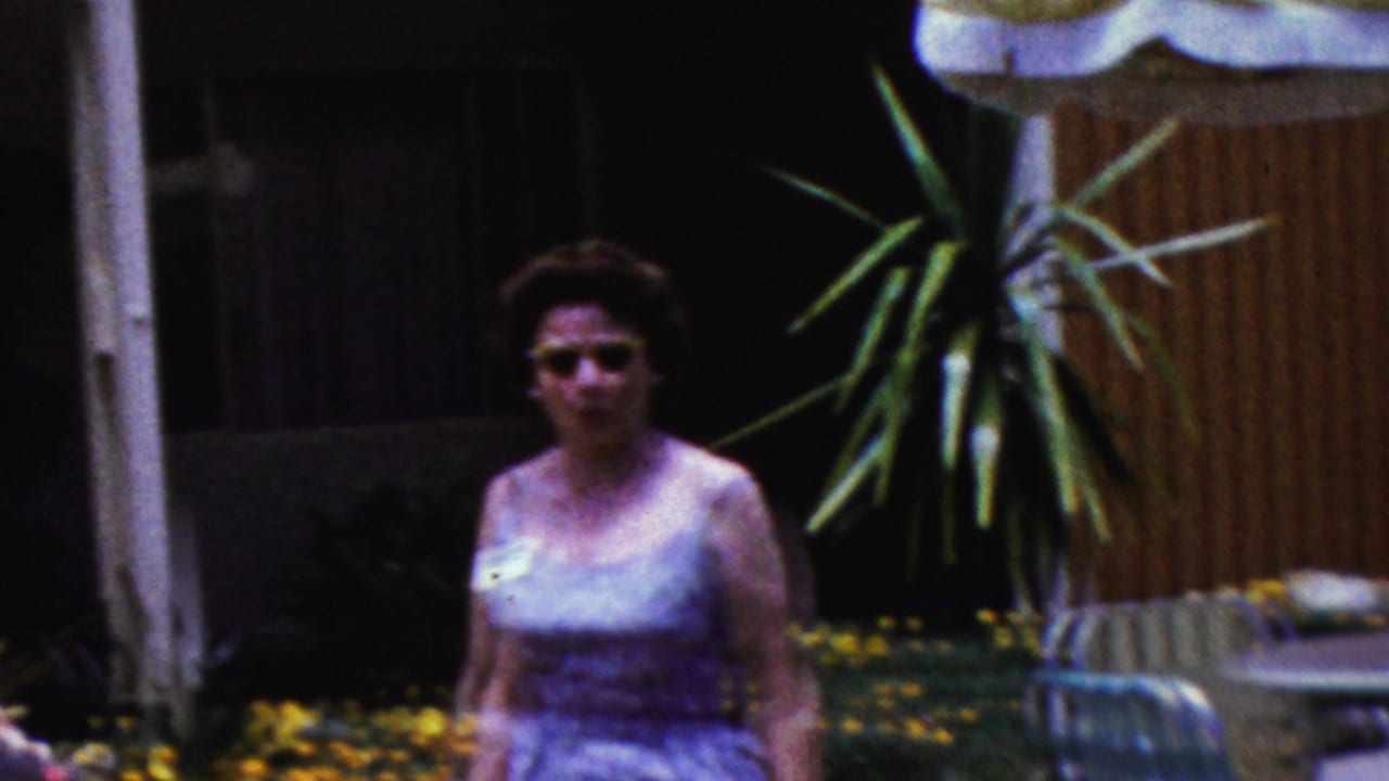 A woman wearing sunglasses and a sleeveless dress stands in a California backyard near plants, flowers, and lounge furniture in 1961 home-movie footage.