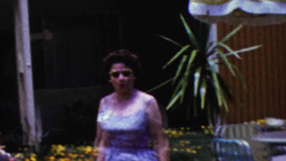 A woman wearing sunglasses and a sleeveless dress stands in a California backyard near plants, flowers, and lounge furniture in 1961 home-movie footage.