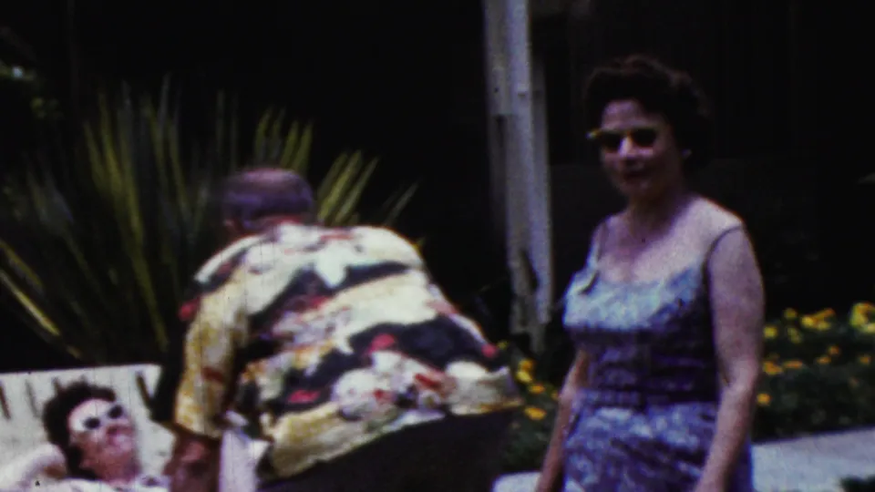 A man in a colorful patterned shirt bends beside a lounge chair as a woman in sunglasses stands nearby in the backyard gathering footage.