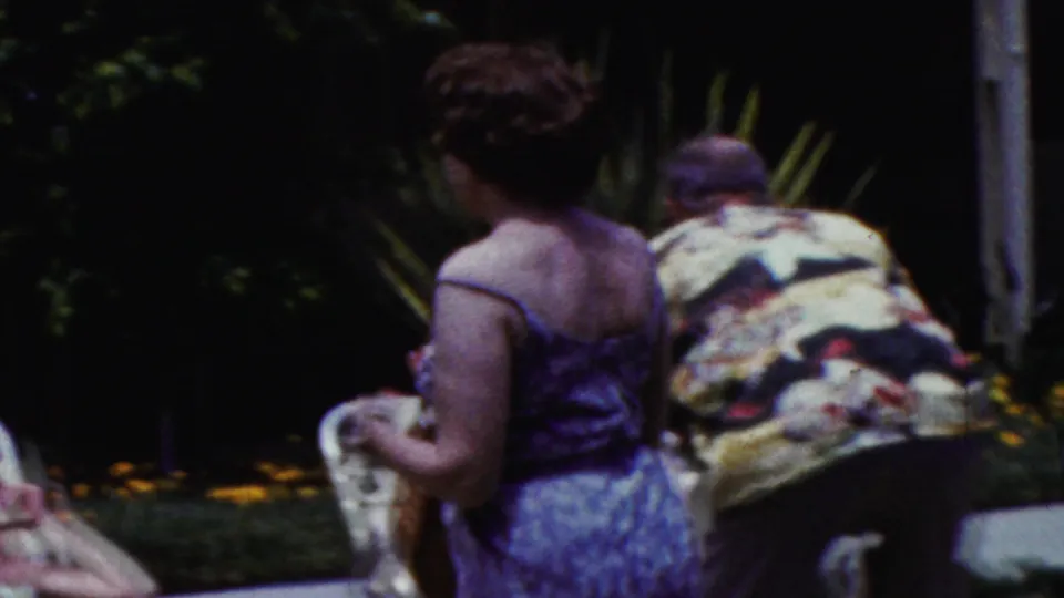 A woman in a sleeveless dress holds a towel or cloth beside a lounge chair while a man in a patterned shirt bends forward in the backyard scene.