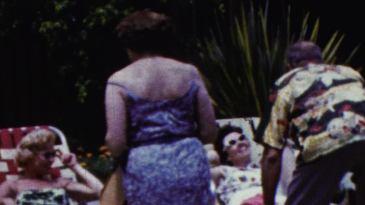 1961 California 8mm frame showing adults around striped lawn chairs, including a woman standing in a purple dress and others reclining in summer clothing.