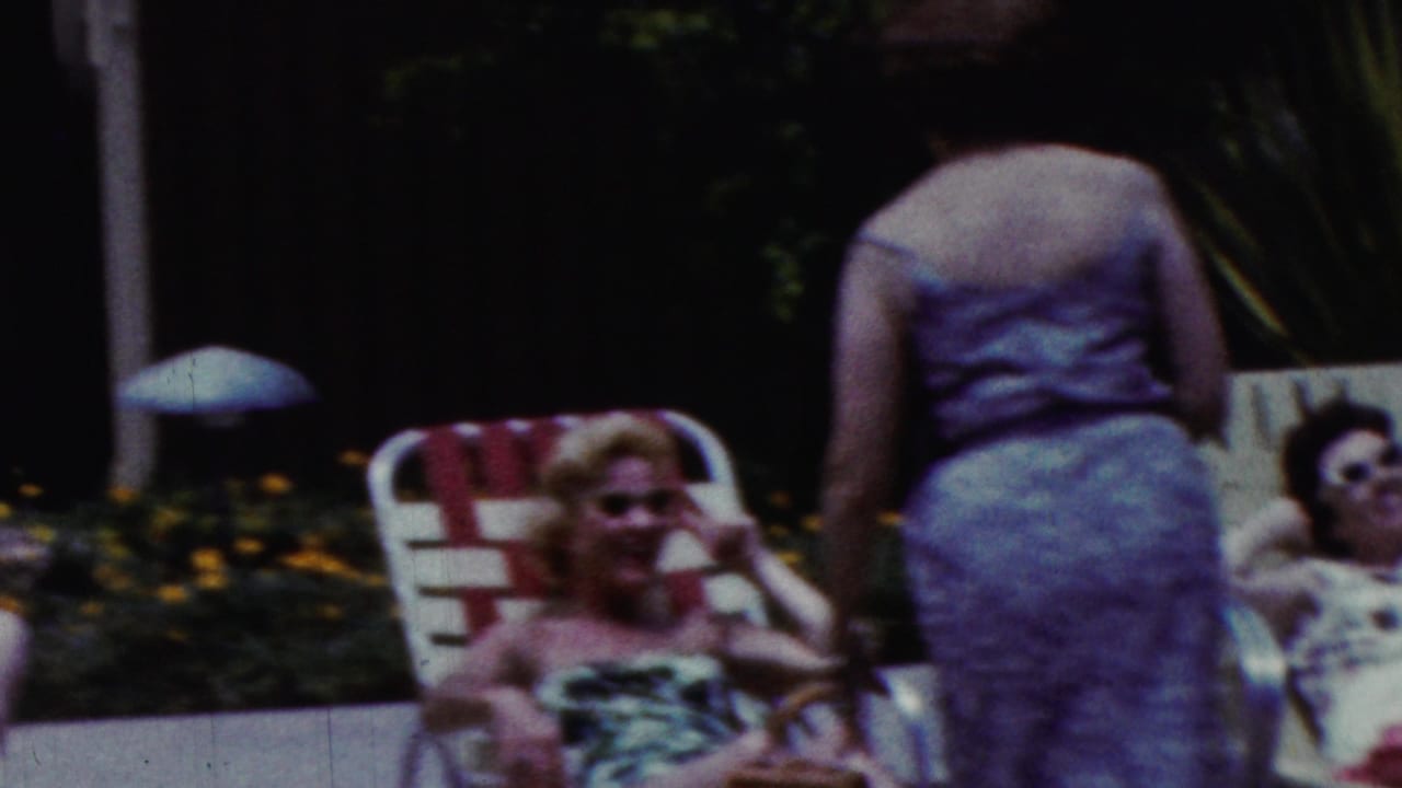 A man bends near a seated woman while another woman stands beside lawn chairs.