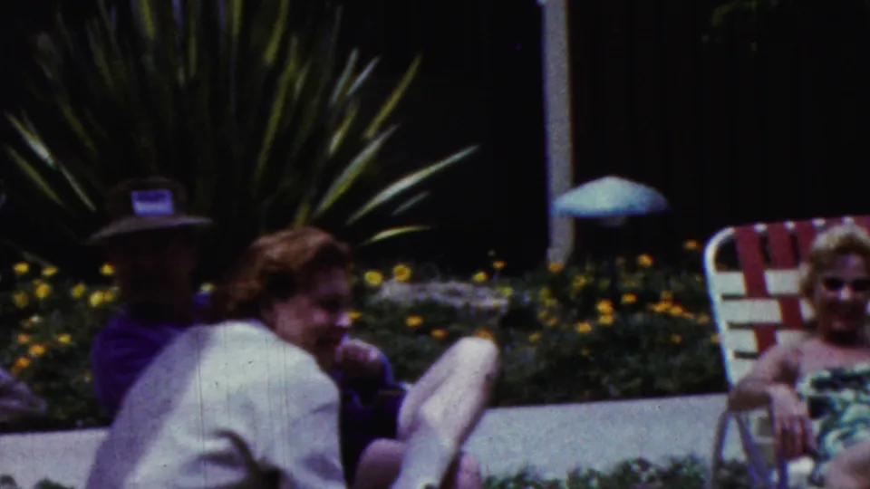 1961 California 8mm frame showing adults seated near a patio table, including a man in a hat and a woman turned toward him, with dense garden planting behind them.