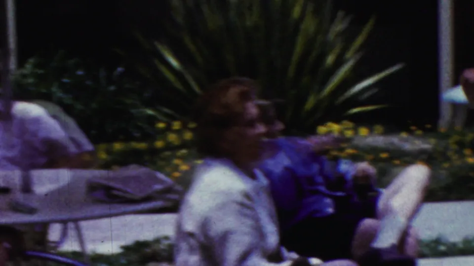 1961 California home-movie frame with a woman seated on a striped lounge chair, a blurred adult passing in front, and another seated guest near garden plants.
