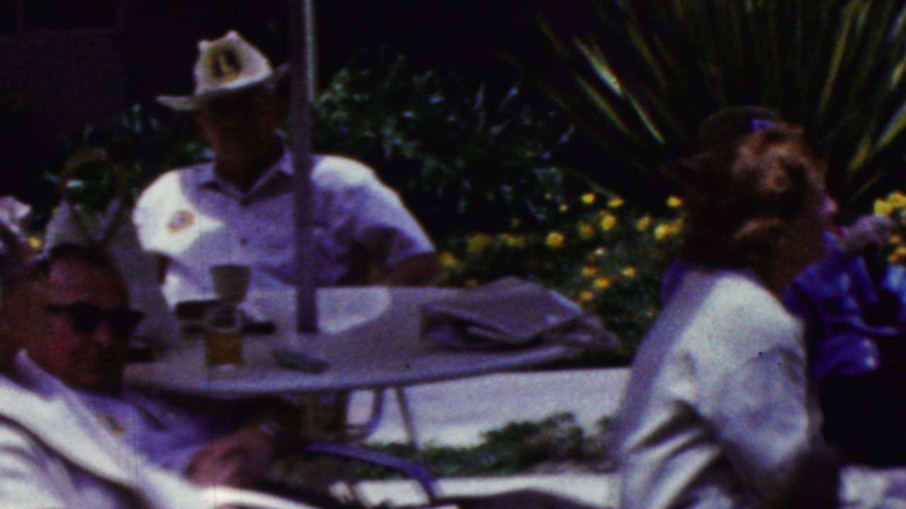 California 1961 home-movie frame showing older adults seated outdoors around a round patio table beneath an umbrella, with sunglasses, light clothing, lounge chairs, and flowering plants visible.